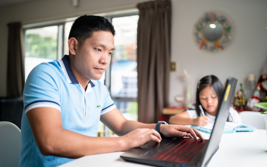 A man sits working on a laptop while his daughter sits doing school work at the same table